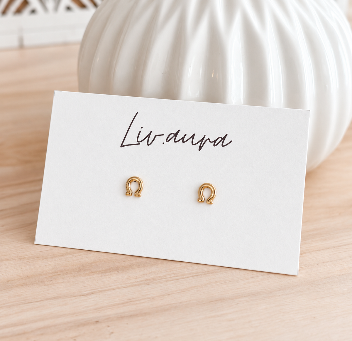 Two small gold hoop earrings displayed on a white card labeled 'Livana' on a light wood surface, with a white ribbed decorative vase in the background.