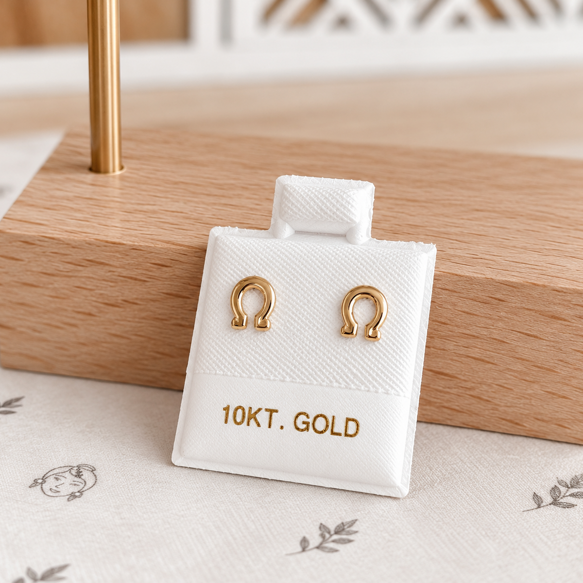 A pair of gold horseshoe-shaped stud earrings displayed on a white jewelry card labeled '10KT. GOLD', set on a light wood block over a cream fabric background.